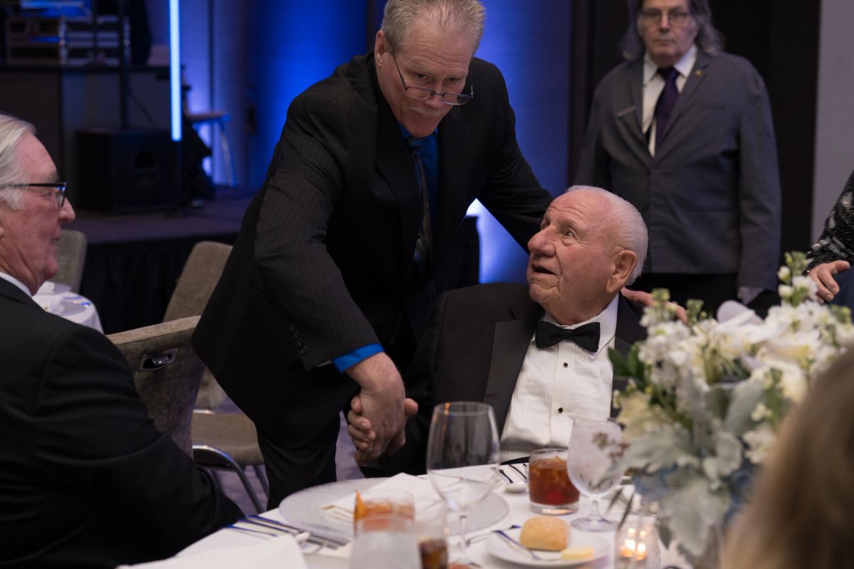 Two men in formal attire shake hands at an elegant dinner table with white flowers and wine glasses.