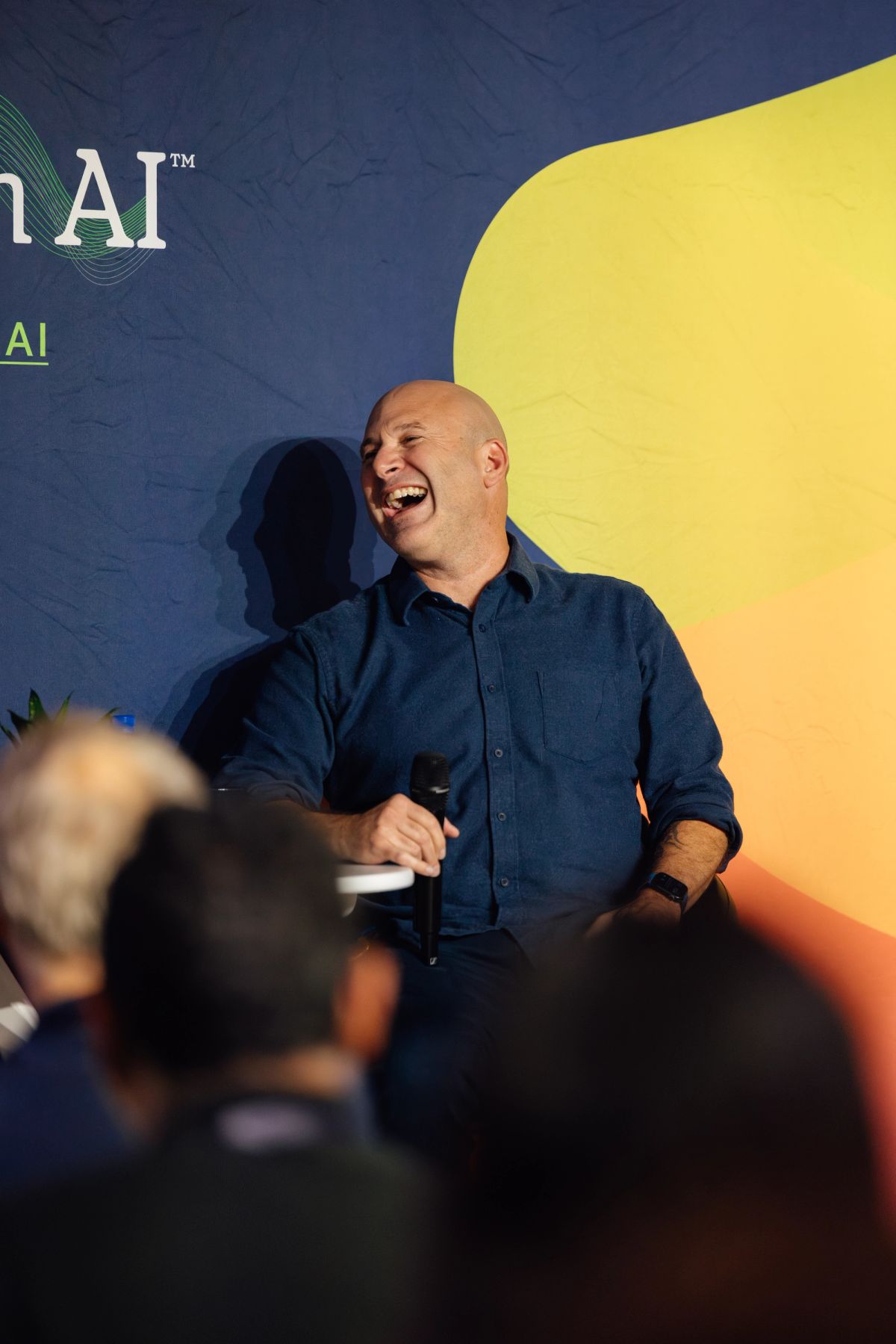 Bald speaker in navy shirt laughing while presenting to audience at conference with yellow and blue backdrop