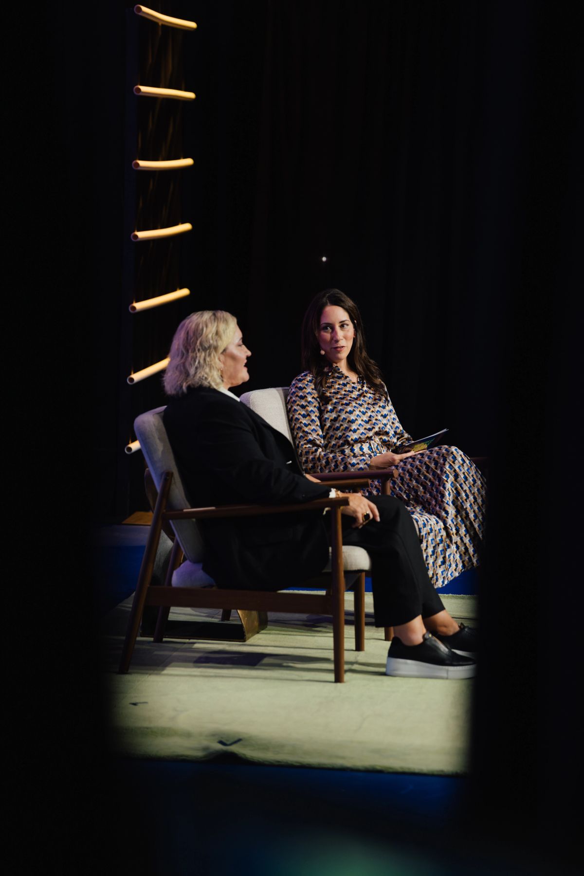 Two women sit in wooden chairs having a conversation on a lit stage with vertical light fixture backdrop.