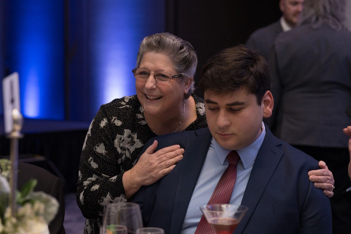Woman in floral jacket embracing young man in suit at formal event with blue lighting and dining tables