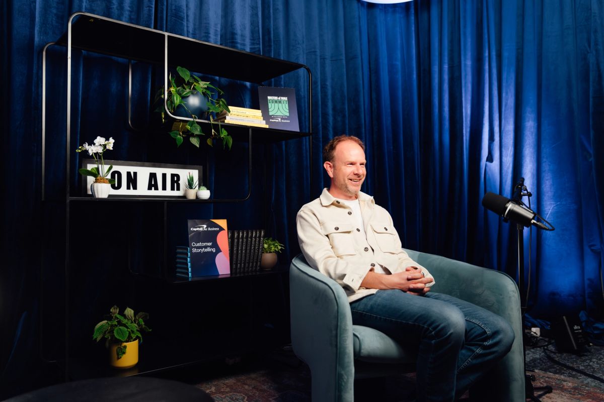Speaker in white shirt sits in blue chair next to bookshelf with "ON AIR" sign against blue curtain backdrop