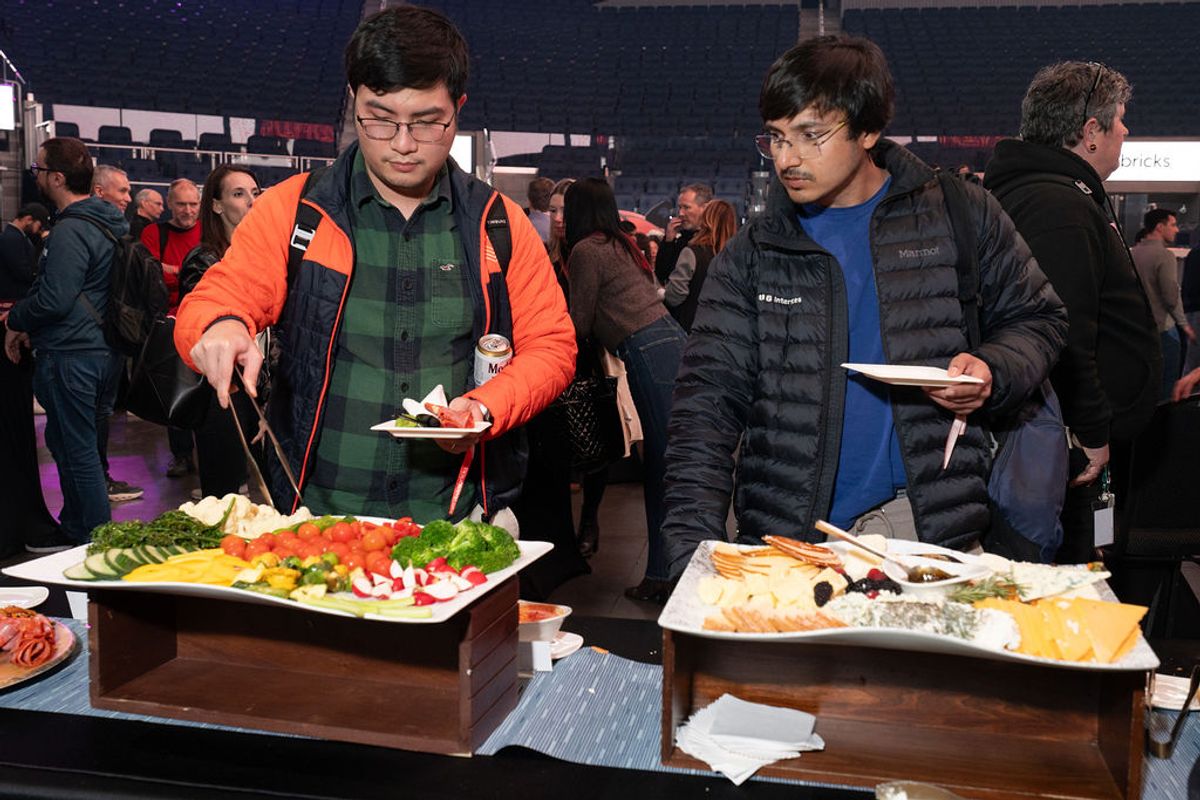 Two attendees serve food from platters at a conference networking event with crowd in background.