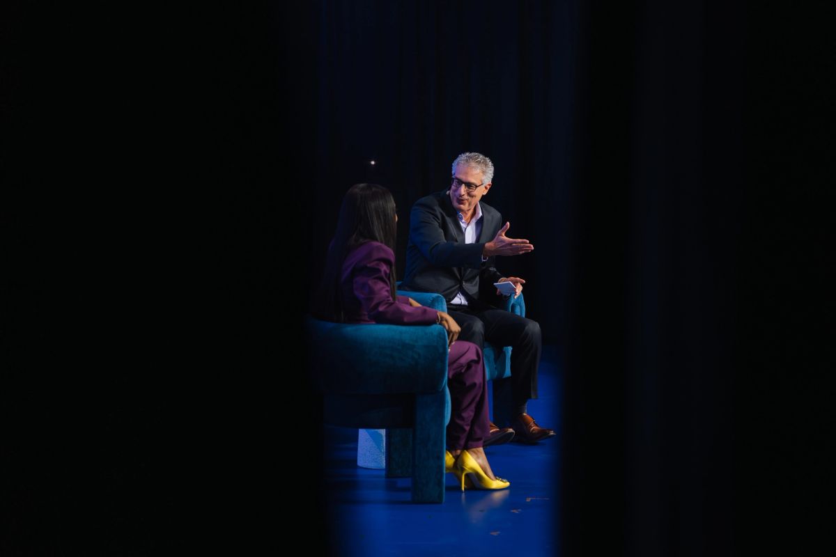 Two people in conversation on stage, one in purple outfit with yellow shoes, one in dark suit, under blue.