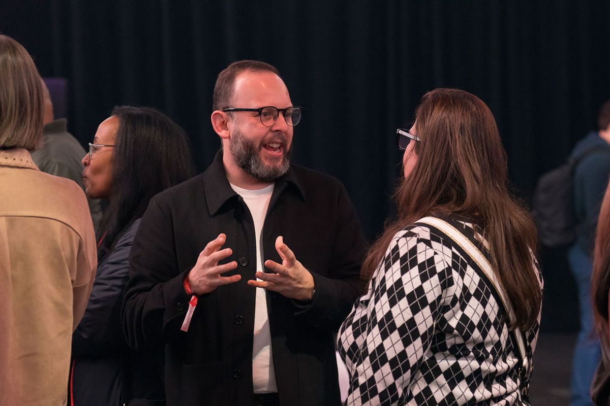 Man in black jacket and glasses speaking with gestures to woman in checkered sweater at networking event