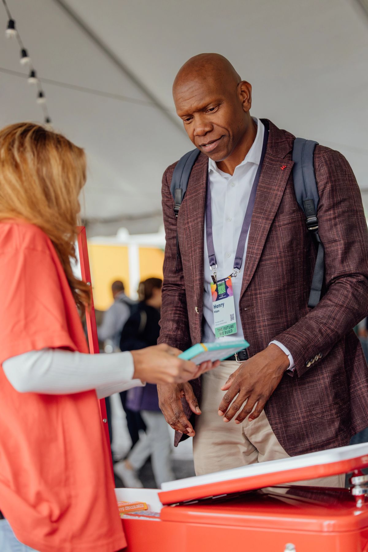 Two professionals shake hands at a conference, man in burgundy blazer with lanyard meeting woman in coral.