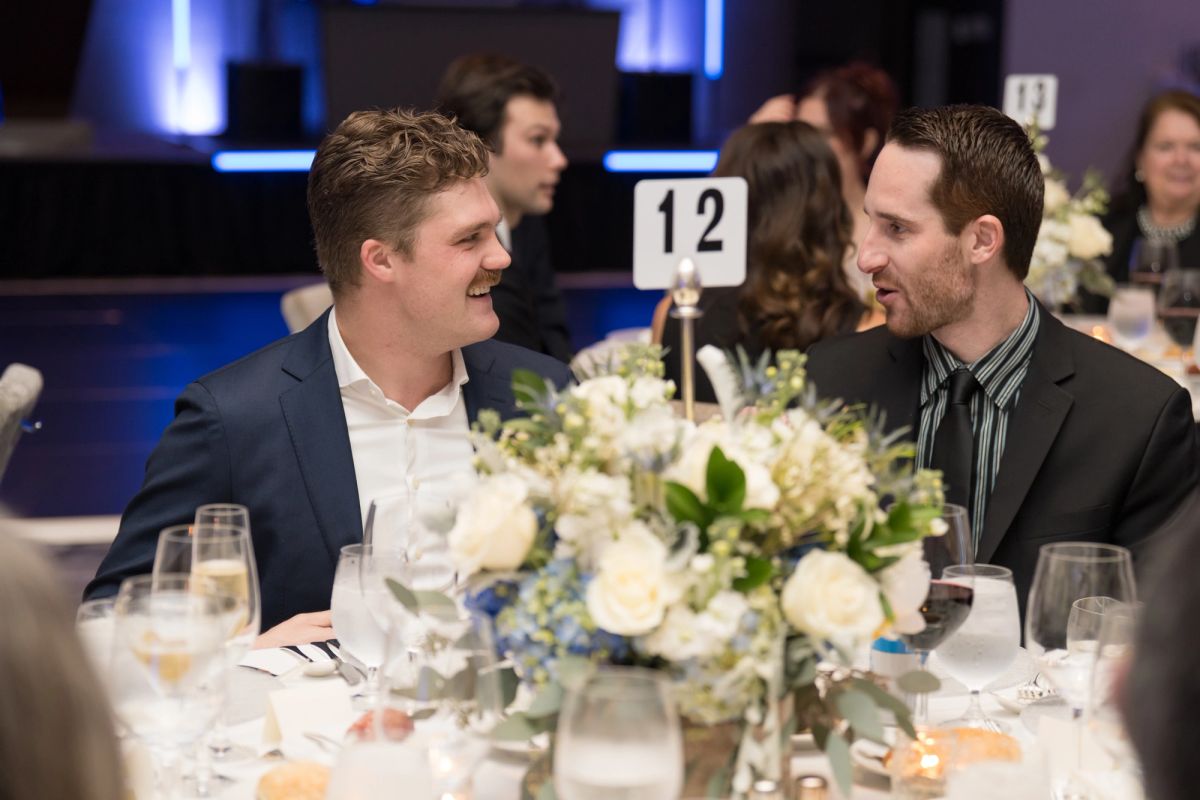 Two men in suits conversing at a formal dinner table with white floral centerpiece and table number 12