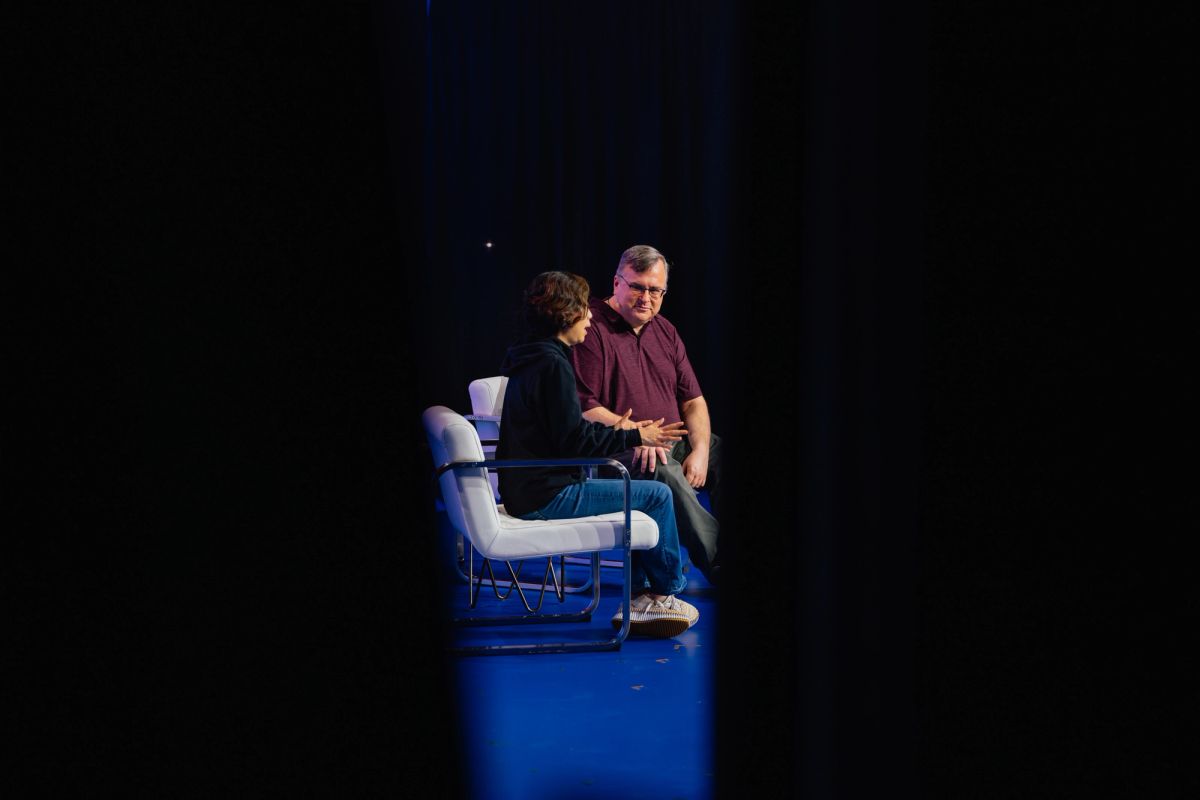 Two people sitting in white chairs on a blue circular platform during a conference presentation