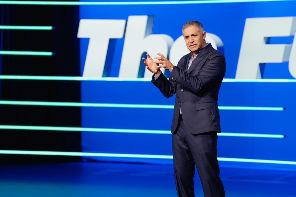 Executive in dark suit gesturing while presenting on stage with blue backdrop at conference venue