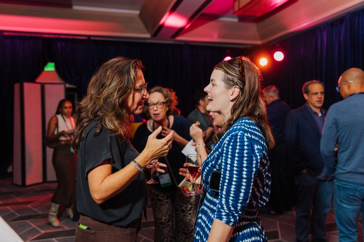 Two women networking and conversing with drinks at an indoor conference event with colorful lighting