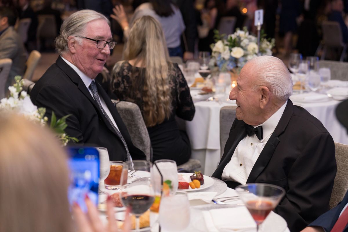 Two men in formal attire having a conversation at a dinner table during an elegant evening event.