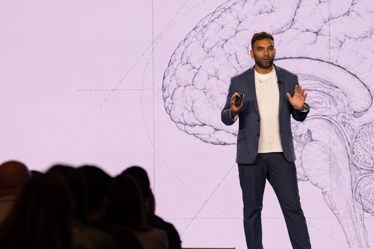 Speaker presenting on stage before large projected brain illustration, gesturing to audience.