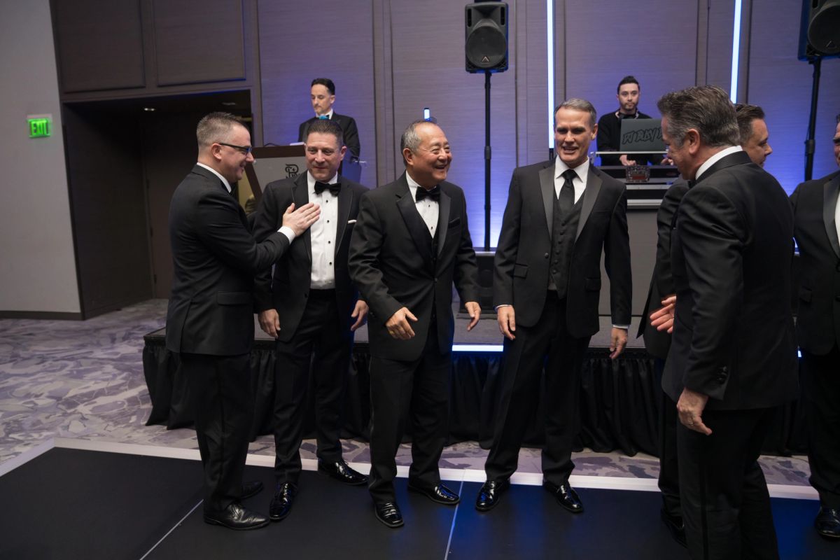 Five men in formal tuxedos gather together at an elegant gala event with purple lighting and DJ booth.