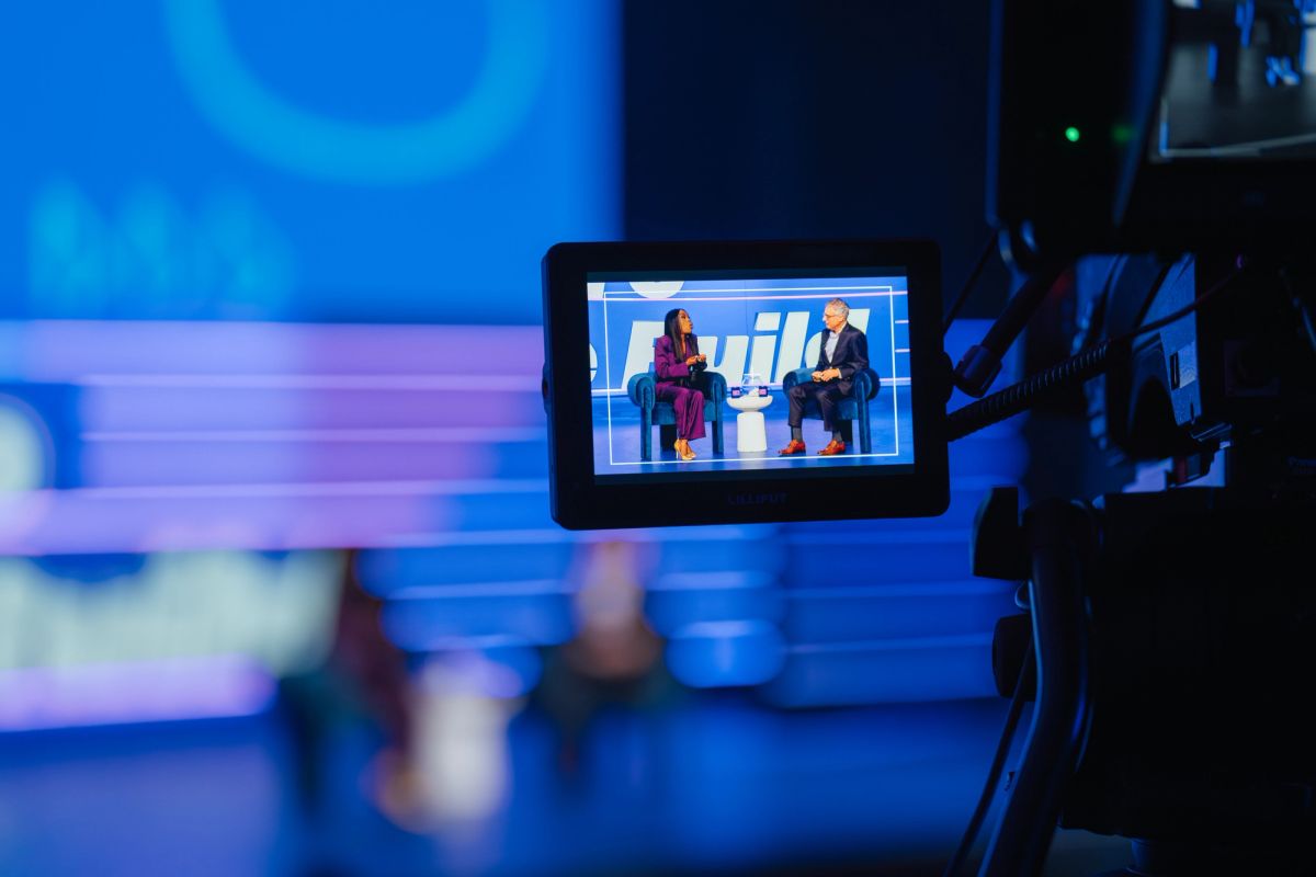 Camera monitor shows two speakers in conversation on stage at conference venue with blue lighting