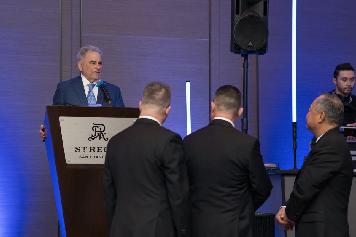 Speaker at podium addressing formally dressed attendees at St. Regis venue with blue lighting