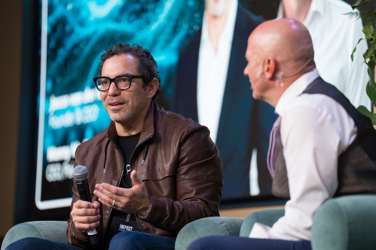 Two speakers seated on stage during a panel discussion, one holding a microphone while speaking to the other.