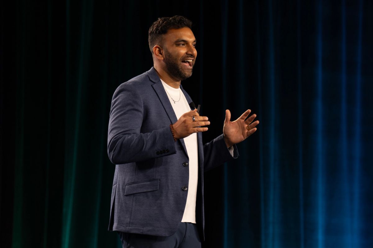 Speaker in navy blazer gestures while presenting on stage before teal curtain backdrop.