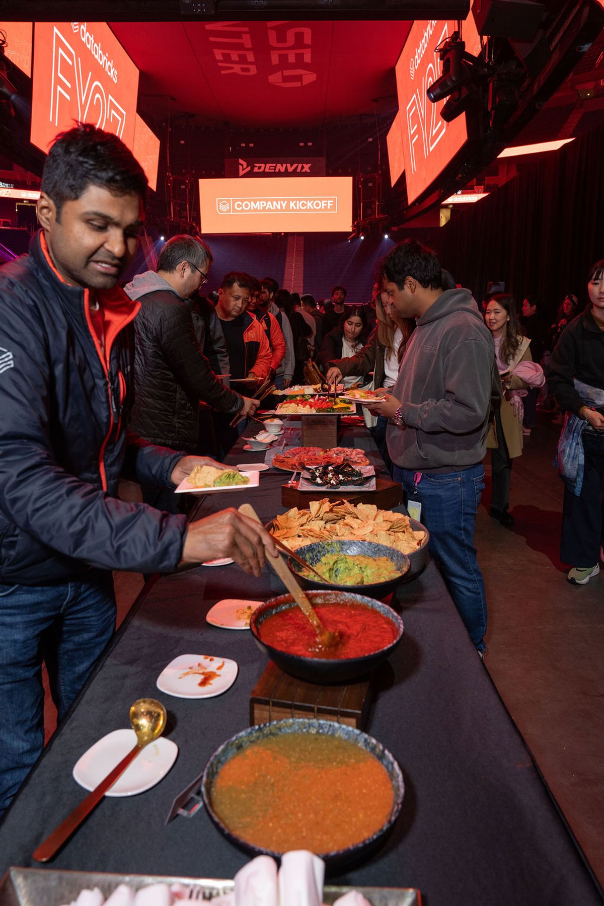 Attendees serve themselves food from large pans at a buffet during a company event with red lighting.