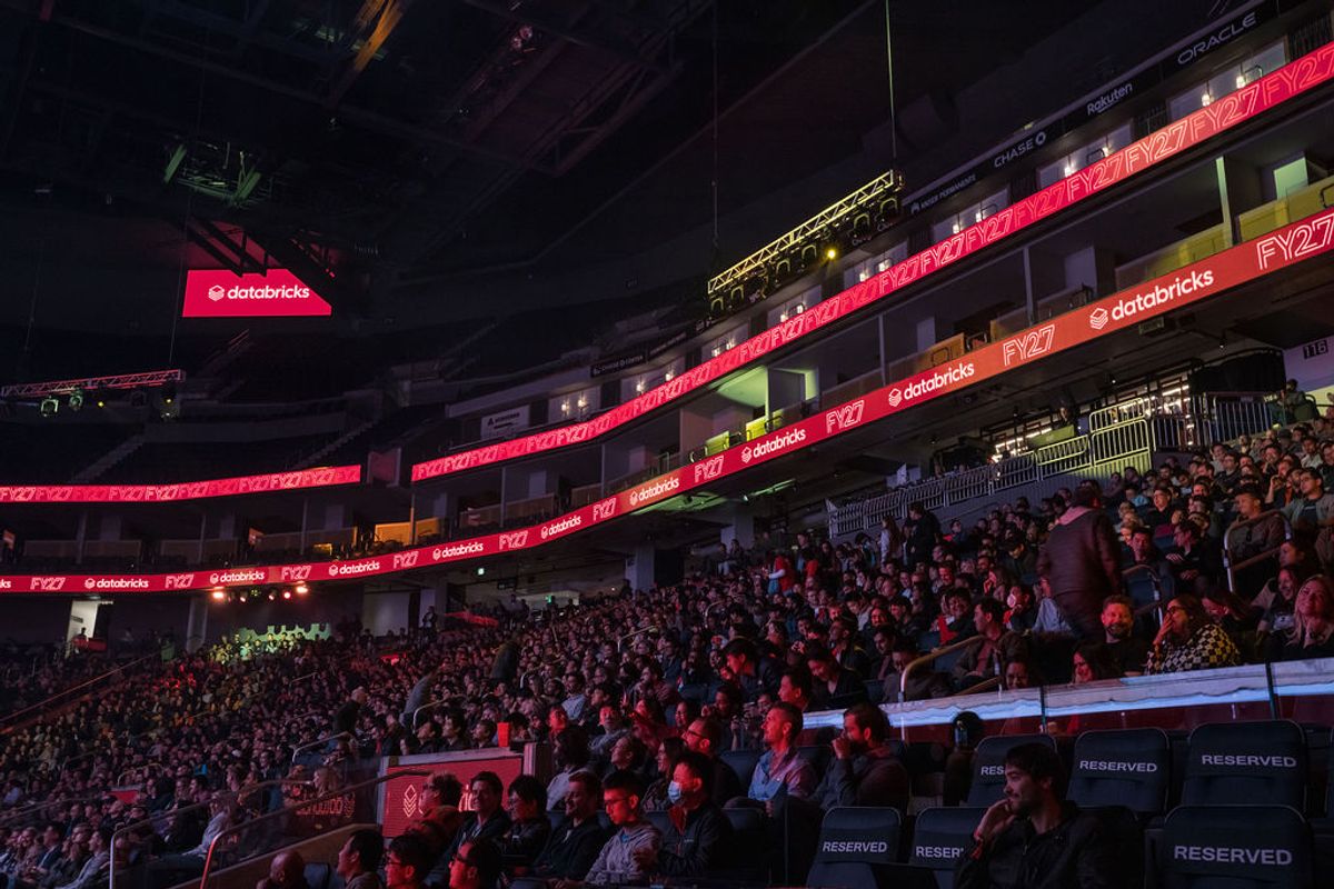 Large crowd of attendees seated in tiered seating at an indoor arena venue with red lighting and digital.