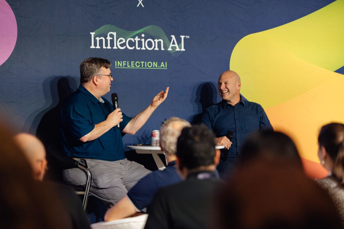 Two speakers on stage in conversation at Inflection AI event, one holding microphone, audience visible below