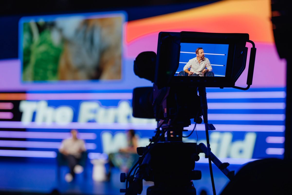 Camera viewfinder showing speaker on stage at conference with colorful backdrop and screens