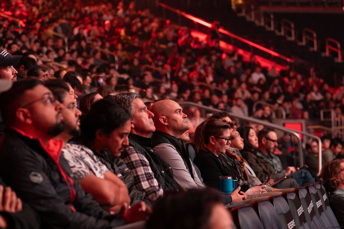 Audience members seated in arena stands watching an event, with red stage lighting illuminating the crowd