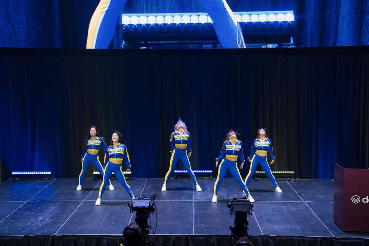 Five cheerleaders in blue and yellow uniforms performing on stage at an indoor venue with blue lighting
