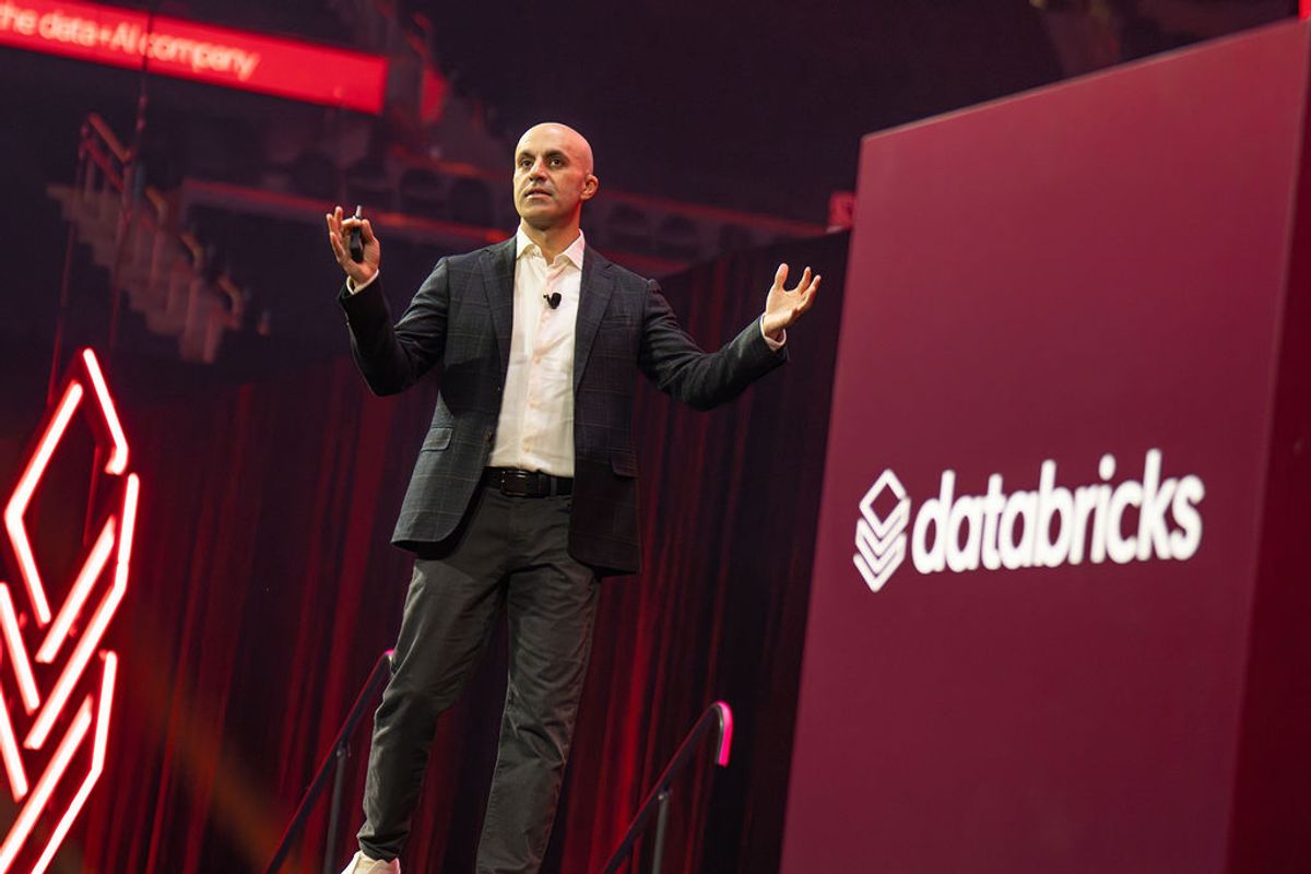 Executive presenting on stage with hands raised, red lighting and Databricks logo visible on backdrop