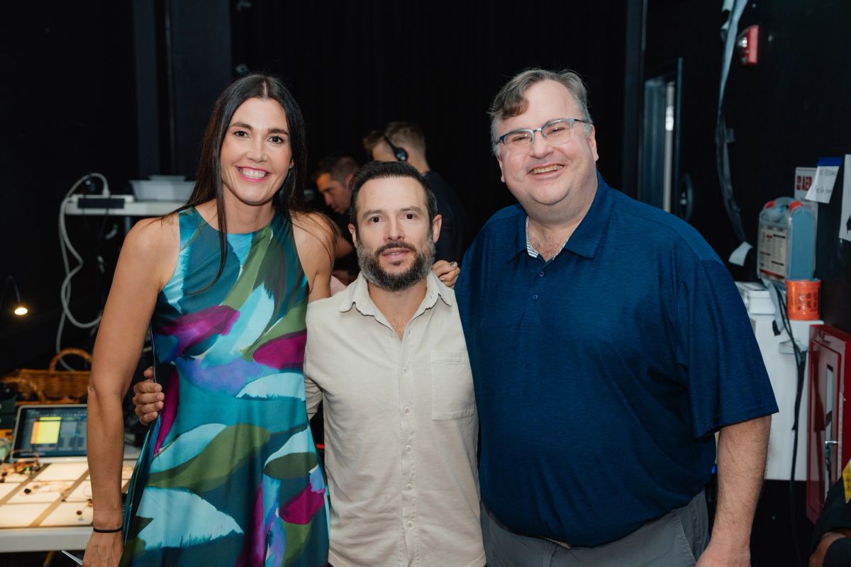 Three people posing together backstage in a theater setting with equipment and cables visible