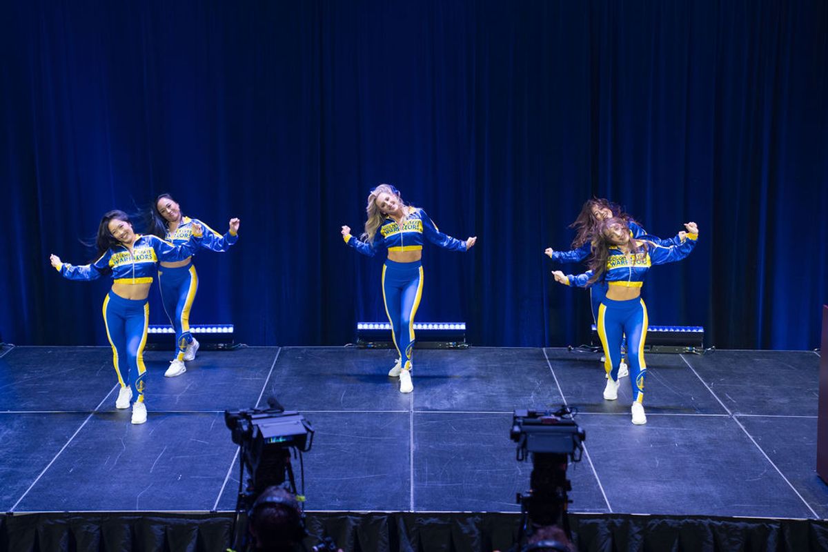 Six cheerleaders in blue and yellow uniforms perform a synchronized routine on stage with blue curtains