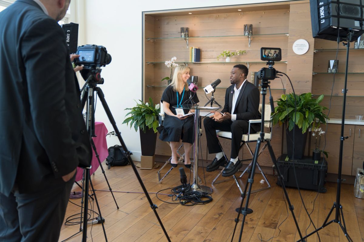 Television interview setup with interviewer and interviewee seated at table, surrounded by cameras and.