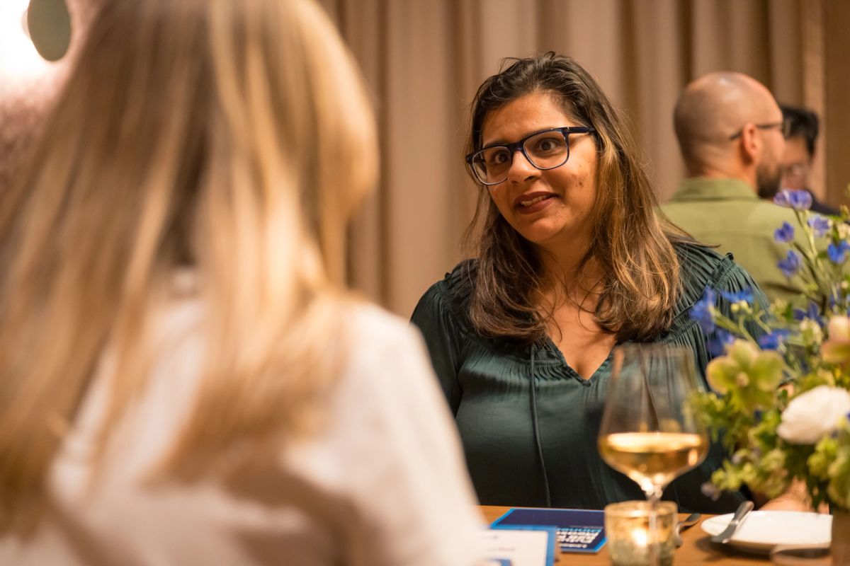 Woman in glasses and dark green top smiling at panel discussion, with wine glass and flowers on table