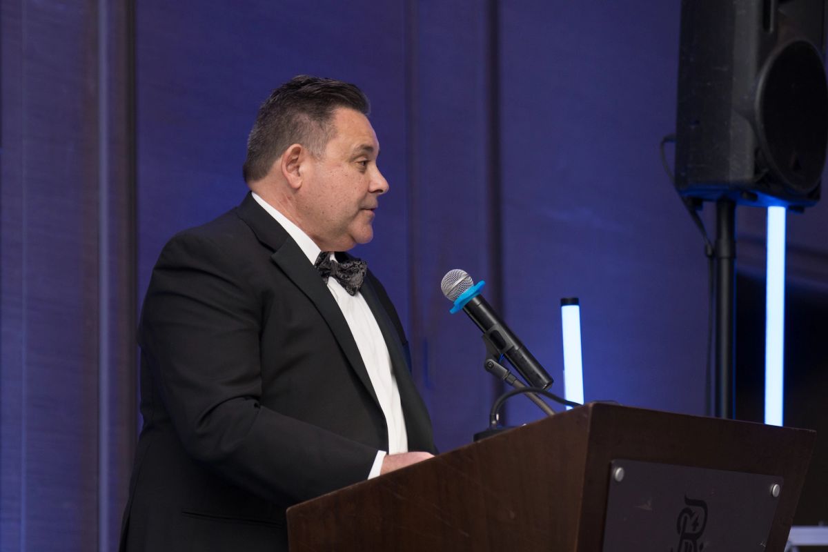 Man in black tuxedo speaking at wooden podium with microphone at formal gala event