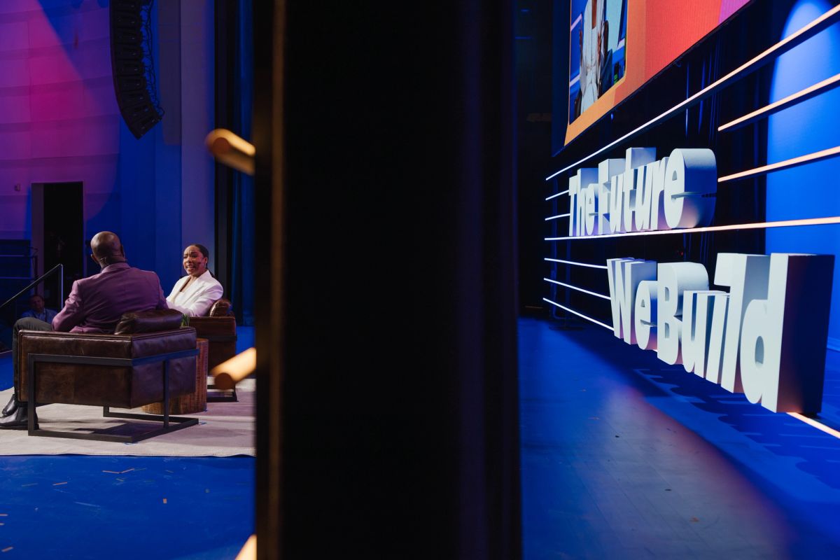 Two speakers sit on stage in chairs during a panel discussion, with blue lighting and event signage visible.