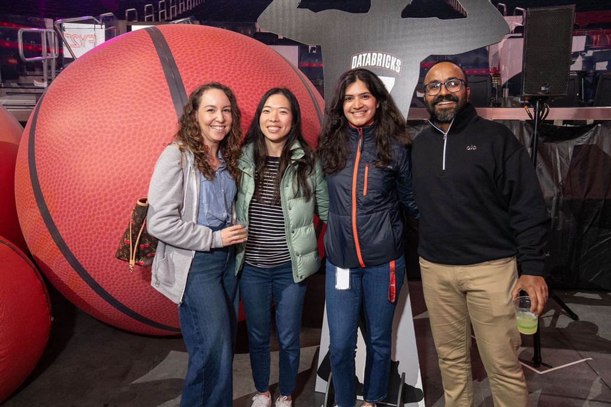 Four people pose together at a basketball arena with a large basketball decoration and Raptors banner visible