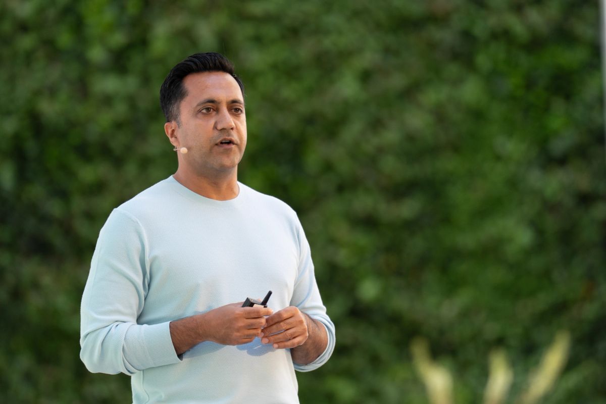 Speaker presenting outdoors, wearing a light blue sweater and lapel mic, holding a clicker.