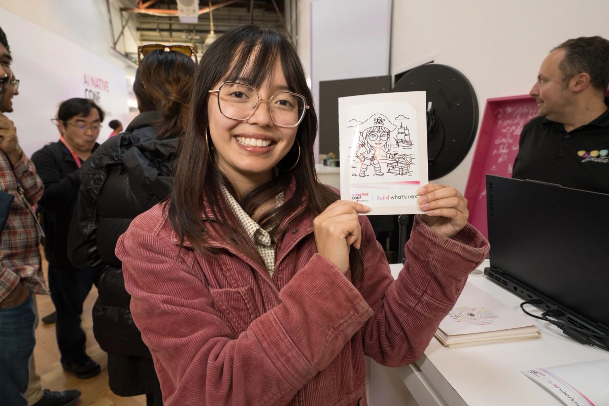 Woman with glasses holds up drawing at busy conference booth with laptop and attendees in background.
