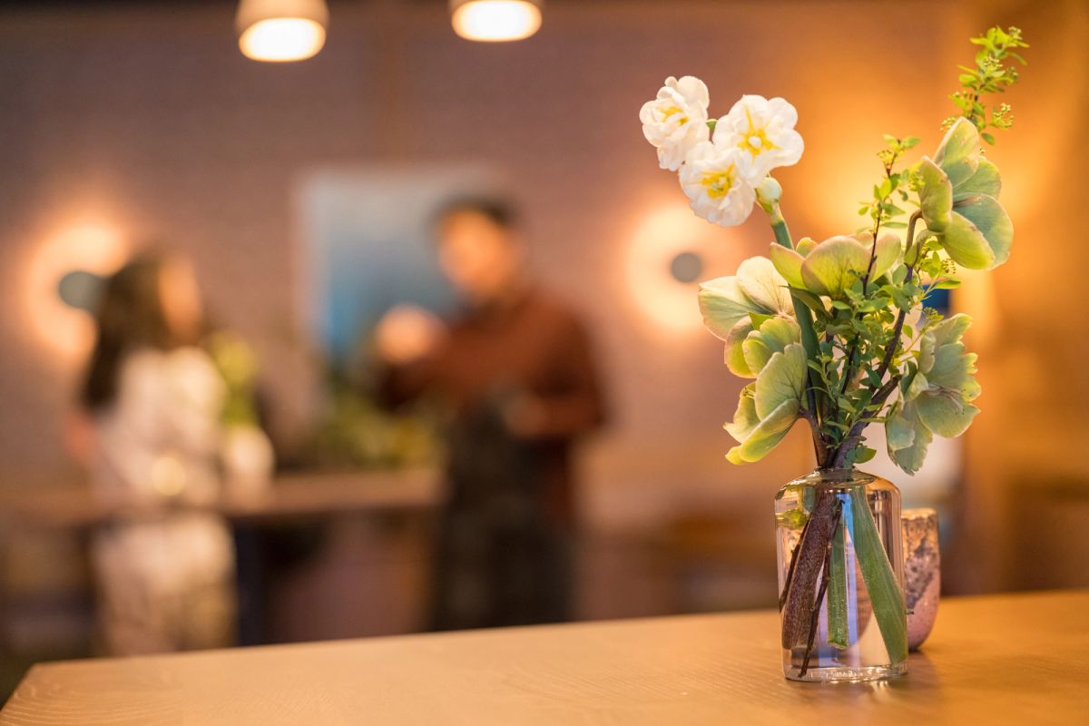 Glass vase with white flowers and green foliage on wooden table, with blurred people in background at event