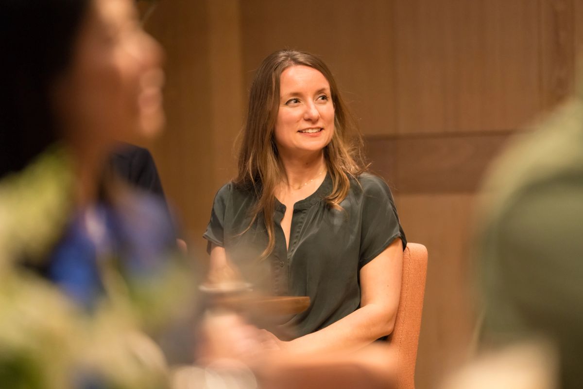 Woman in green shirt sitting in orange chair during panel discussion, with blurred audience members visible
