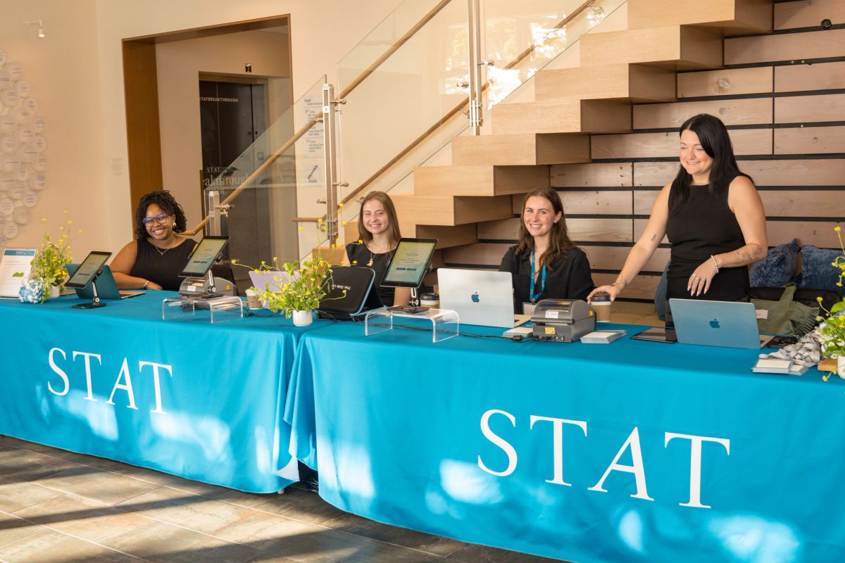 Four women at registration table with blue STAT tablecloth, laptops and materials in modern lobby