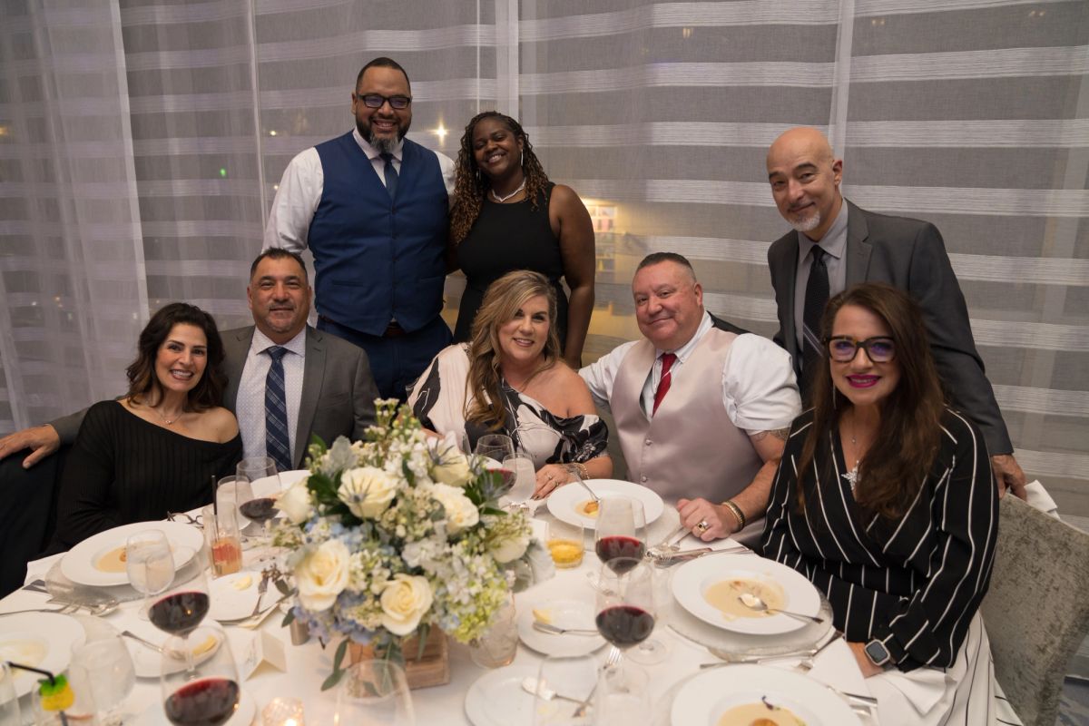 Seven people in formal attire pose together at a dinner table with white flowers and wine glasses