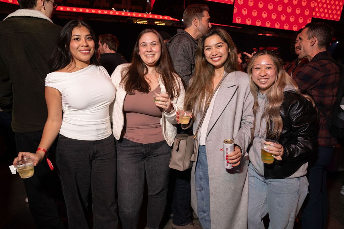 Four women pose together holding drinks at an indoor event with red lighting and other attendees nearby.