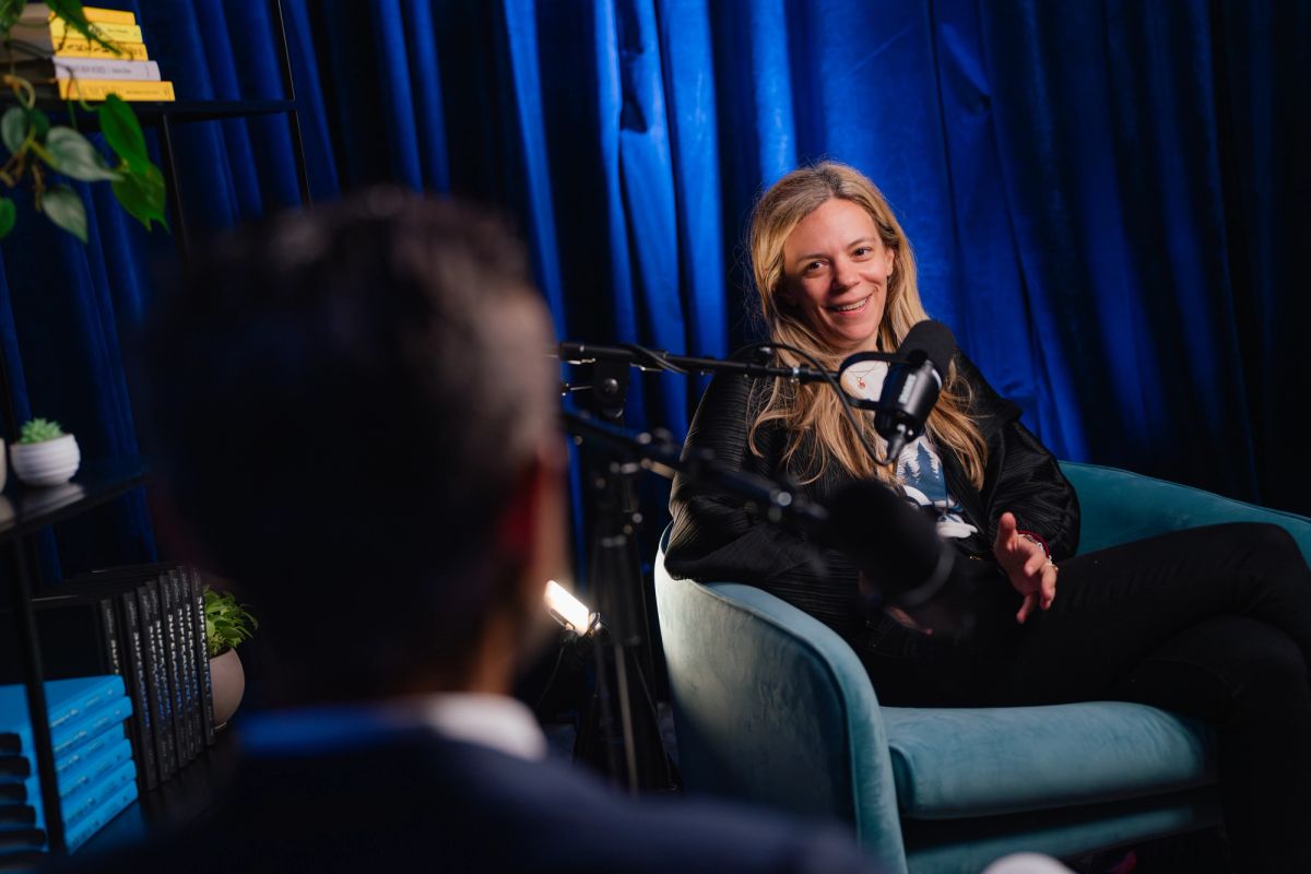 Woman in black outfit sits in blue chair speaking into microphone during interview, with blurred interviewer.