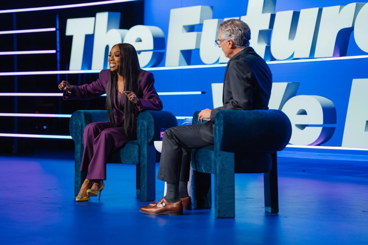 Two people sitting in blue chairs on stage having a conversation, with "The Future" visible on blue backdrop
