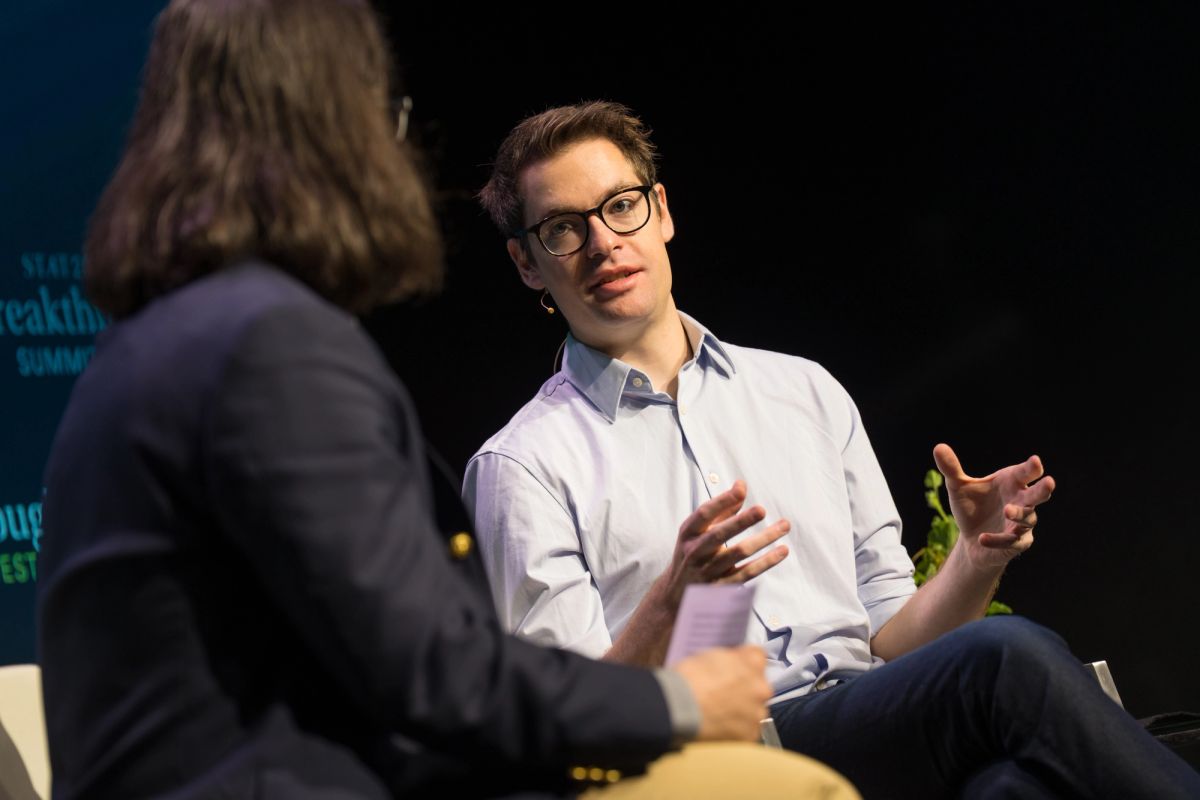 Speaker in light blue shirt and glasses gestures while speaking to interviewer in dark suit on stage.
