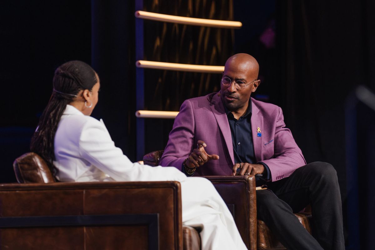 Two people in conversation on stage, one in white outfit and one in purple blazer, seated in wooden chairs.