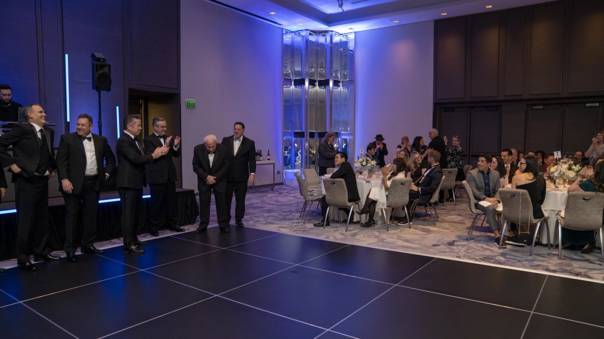 Group of men in formal attire standing on dark tiled floor addressing seated dinner guests in an elegant.