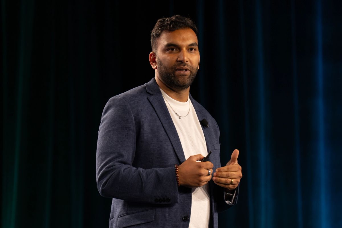 Speaker presenting on stage, wearing blue blazer and white shirt, holding a clicker.