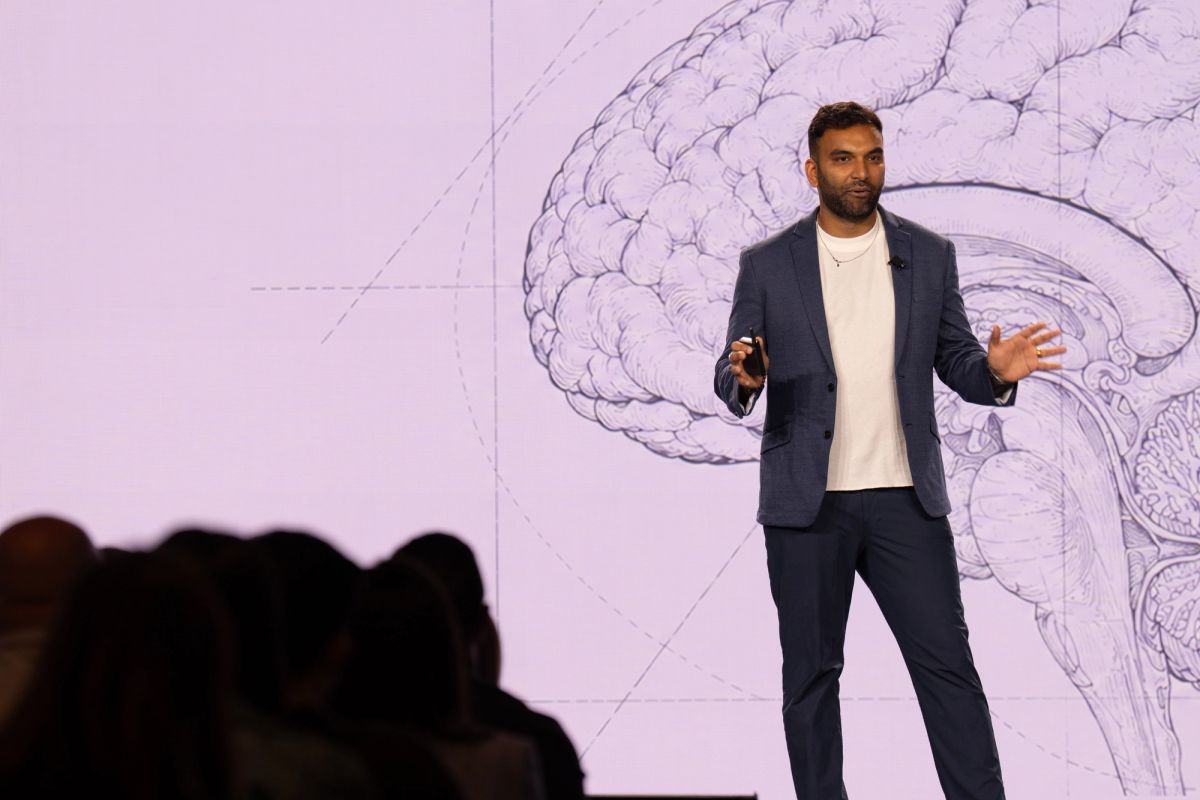 Speaker presenting on stage before large projected brain illustration, gesturing to audience.