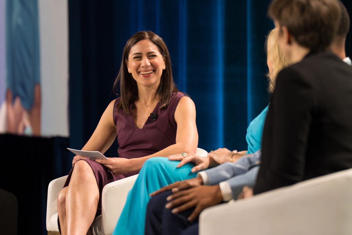 Smiling panelist in burgundy dress holds tablet during conference panel discussion on stage.