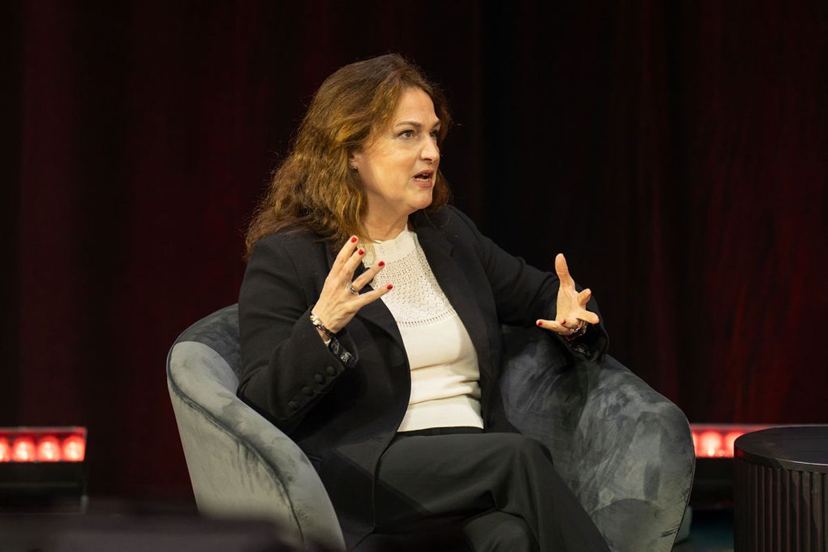 Speaker in dark suit gesturing while seated in gray chair during conference presentation